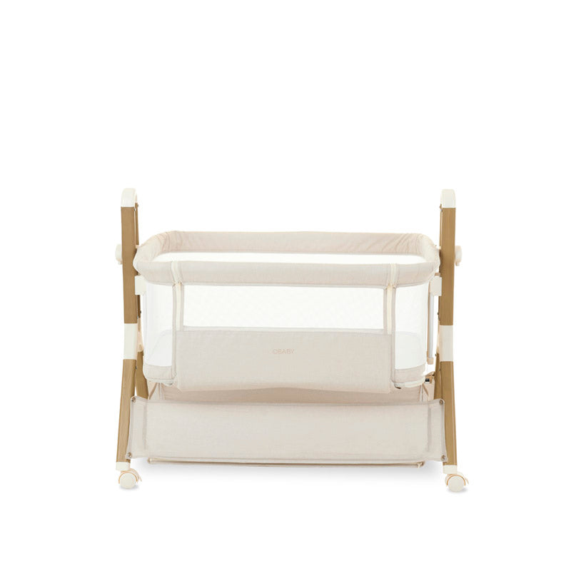 bedside crib, with fabric oatmeal, mesh sides, wood effect frame, crib on lowest setting, storage basket underneath crib for parent easy access. 