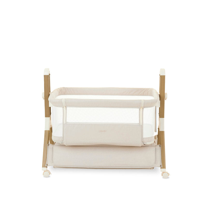 Bedside crib on its lowest setting, with oatmeal fabric, wood effect frame, storage basket underneath crib. Caster wheels to help move the crib into position 
